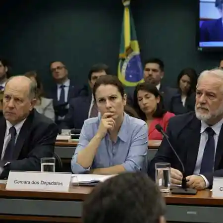 Cena de audiência pública em sala de comissão legislativa mostrando especialistas e parlamentares atentos, com expressões faciais sutis indicando concentração, dúvida e avaliação crítica.
