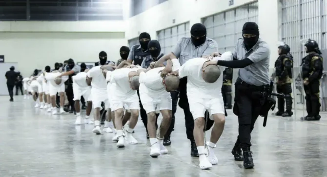 Presos em celas do CECOT, em El Salvador, observados por policial, em imagem sobre encarceramento, controle prisional e segurança pública.